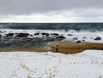 Boy walking at snow covered beach against sky