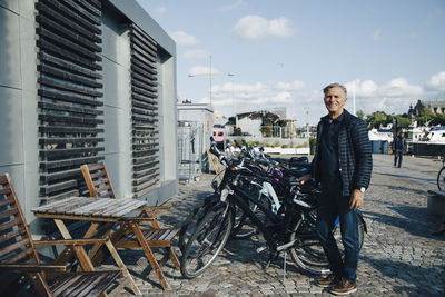 Full length portrait of smiling senior man standing by bicycle at parking area in city
