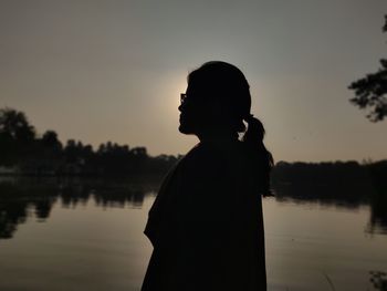 Side view of silhouette woman standing by lake against sky during sunset