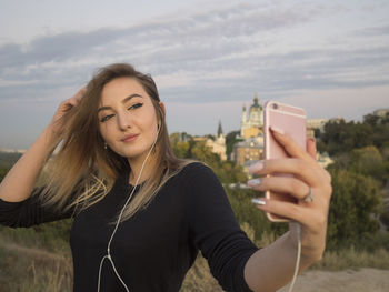 Woman listening music while taking selfie through mobile phone against sky