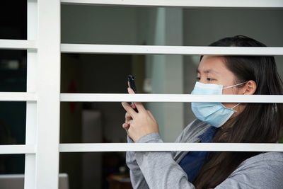 Side view of woman wearing mask using mobile while sitting by window