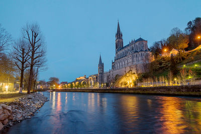 Illuminated buildings by river against sky in city at dusk