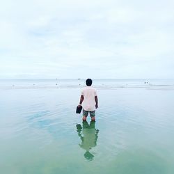 Rear view of man holding footwear while standing in sea against sky