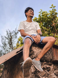 Low angle view of happy young man sitting on retaining wall against sky