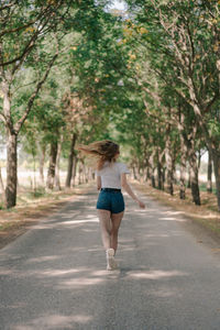 Back view of anonymous female jogger in sports clothes running on pathway between trees during workout in daytime