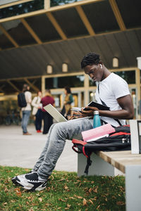 African male student reading book while sitting on bench in university campus