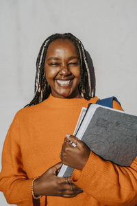 Portrait of cheerful young woman with braided hairstyle holding book and file against gray wall on sunny day