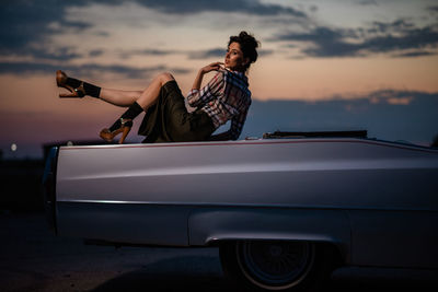 Portrait of woman sitting on car against sky during sunset