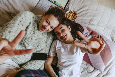 Portrait of happy female friends showing peace signs while lying on bed at home