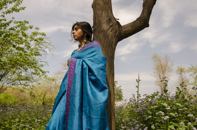 Young woman looking away while standing by tree against sky