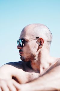 Shirtless mid adult man wearing sunglasses while sitting at beach against clear sky during sunny day