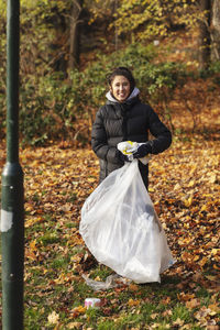 Portrait of smiling female environmentalist with plastic waste standing in park during autumn