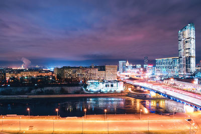 Illuminated city by river against sky at dusk