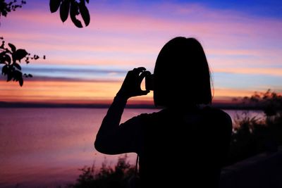 Silhouette woman photographing sea with smart phone against sky during sunset