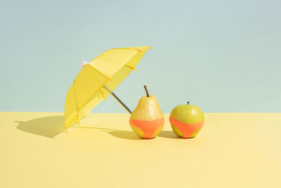 Wet apple and a pear in monokini next to an umbrella isolated on a blue and yellow background.