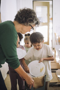 Children throwing leftovers in garbage can held by teacher at kindergarten