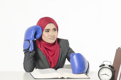 Thoughtful businesswoman wearing boxing gloves while sitting at desk against white background
