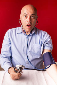 Portrait of shocked man checking blood pressure at table against red wall