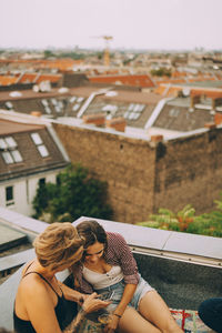 High angle view of female friends looking at mobile phone while sitting on terrace in city during rooftop party