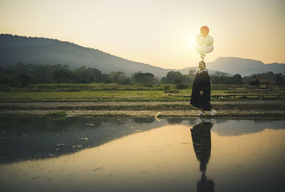 Side view of woman with balloons standing by lake on field against mountain during sunset
