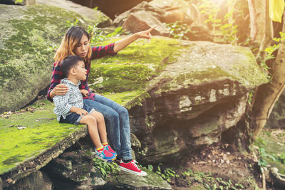 Full length of mother pointing while sitting by son on mossy cliff