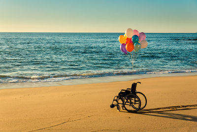 Colorful helium balloons on empty wheelchair at beach against clear blue sky