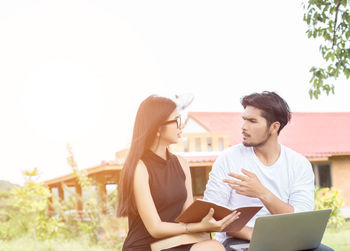 Young man using laptop by woman sitting with book against sky