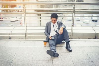 Thoughtful businessman using phone while sitting on elevated walkway in city 