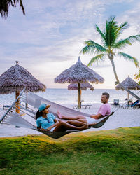 People relaxing on deck chairs by palm trees against sky