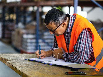 Side view adult worker wearing uniform writing on clipboard while working in spacious storehouse
