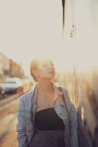 Portrait of young woman standing by wall against sky at sunset
