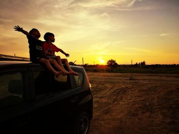 Friends sitting on car roof against sky during sunset