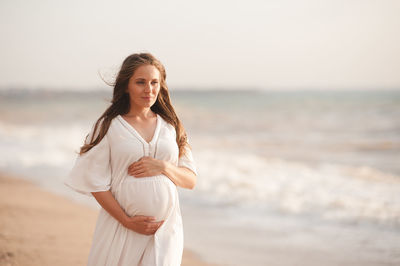 Smiling pregnant woman wear white dress hol tummy walk at beach over sea nature background outdoors