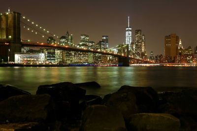 Illuminated bridge over river by buildings against sky at night