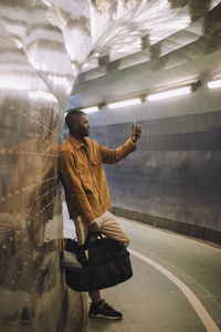 Full length of young man carrying bag while taking selfie through mobile phone in subway tunnel