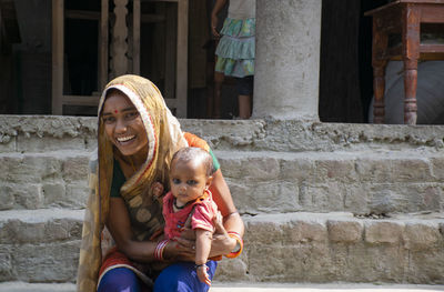 Portrait of smiling young mother and daughter sitting against brick wall in rural india 