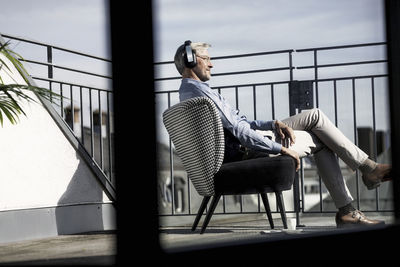 Grey-haired man relaxing in chair on balcony listening to music