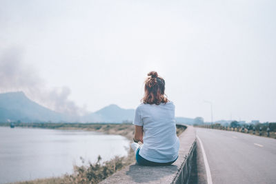 Rear view of woman sitting on retaining wall at lake against sky