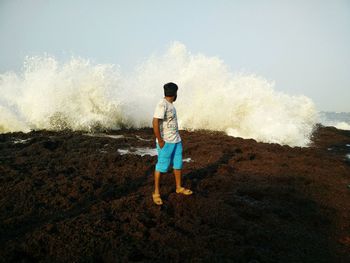 Man with hands in pockets looking at waves splashing on rocks against clear sky