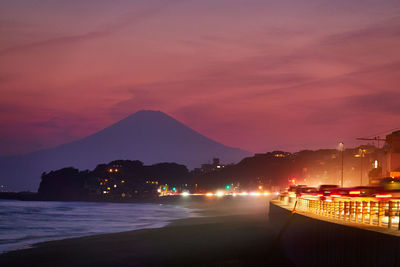 Illuminated city by sea against sky at sunset