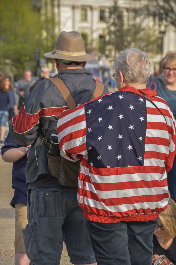 REAR VIEW OF PEOPLE WALKING IN FRONT OF FLAGS