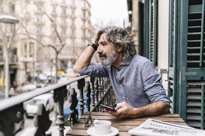 Thoughtful senior man with smart phone sitting at table in balcony at house