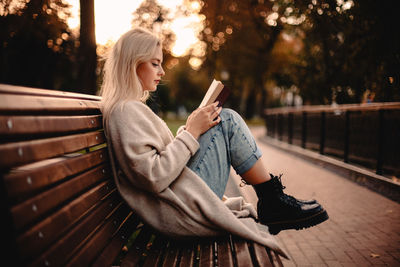 Teenage girl reading book sitting on bench in park during autumn