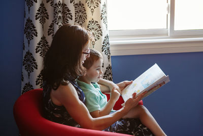 Mother with son boy sitting in armchair at home and reading book together. child kid early education 