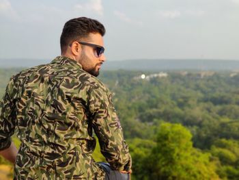 Rear view of young man wearing sunglasses sitting on mountain against sky