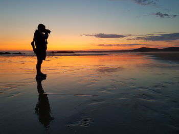 Silhouette man photographing while standing at beach against sky during sunset