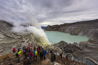 People hiking at volcanic mountains by lake against cloudy sky