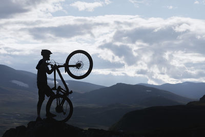 Silhouette cyclist standing with bicycle on mountain peak against sky