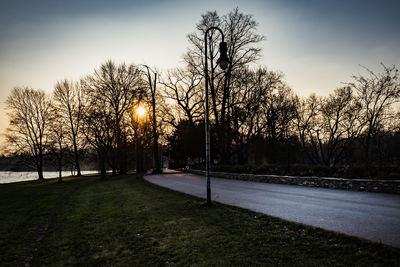 Road by bare trees against sky during sunset