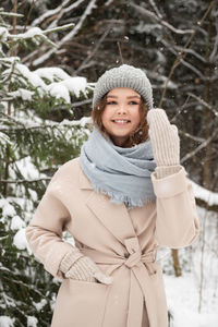 A cute girl is standing under the snow and smiling in the forest in mittens and coats